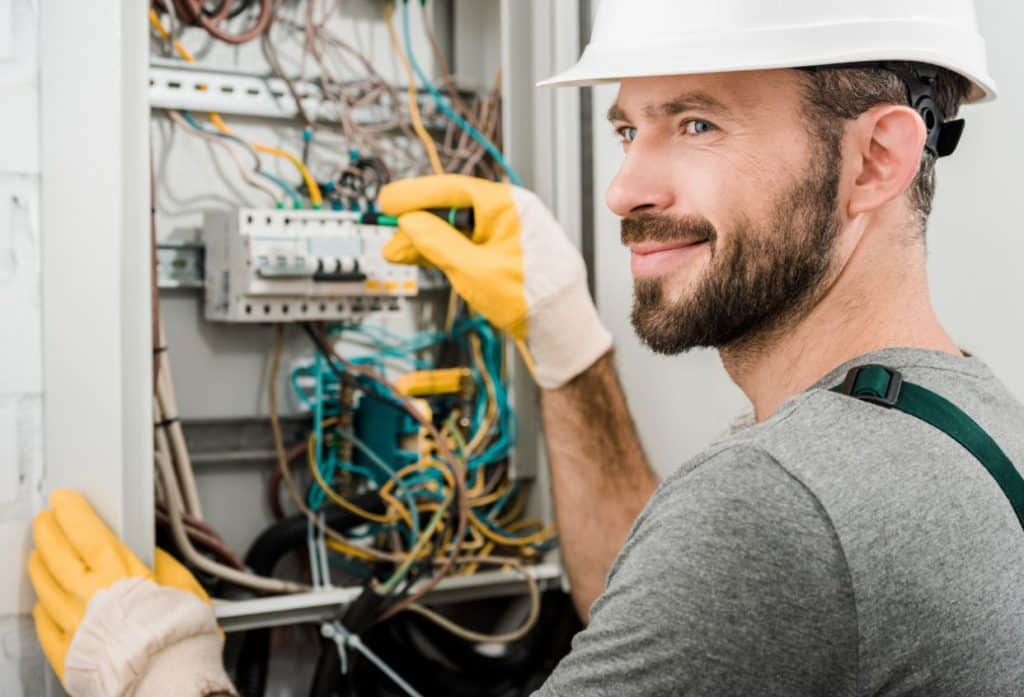 Electrician wearing a hard hat and protective gloves inspecting an open electrical panel filled with wiring, circuit breakers, and electrical components.